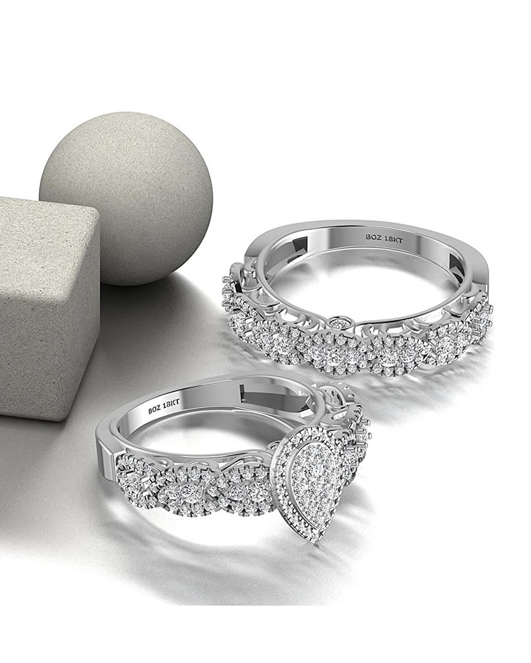Two 18K gold rings with pear cut diamonds, one larger and one smaller, displayed on a reflective surface with a grey sphere in the background.