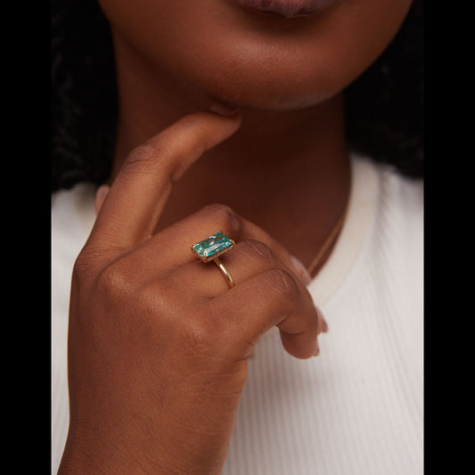 Close-up of a hand wearing a gold ring with a green gemstone, against a neutral background.