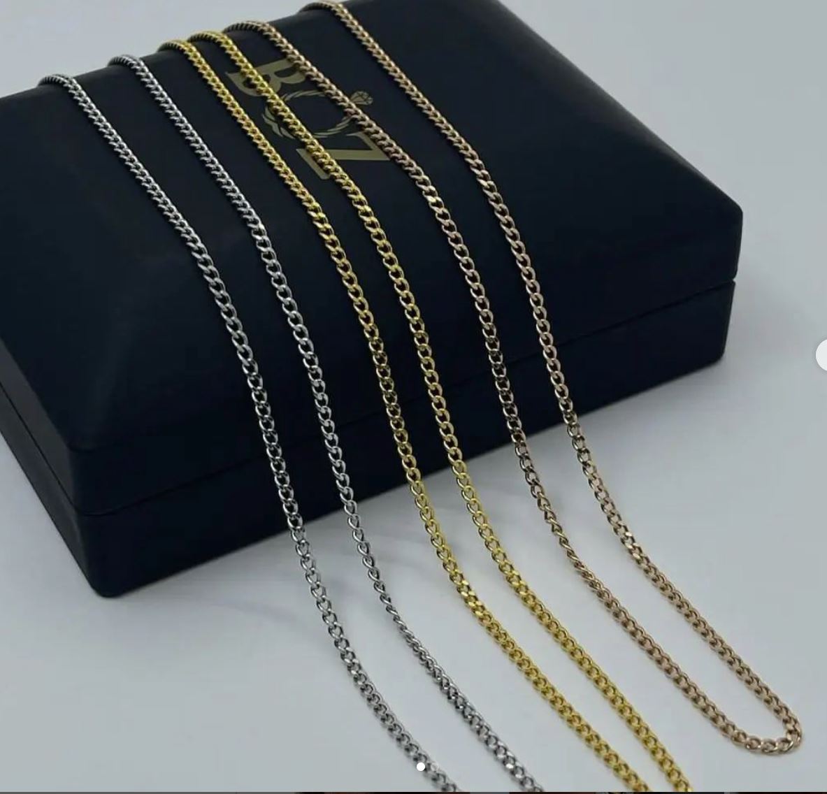 A collection of thin cable necklaces in 18K gold displayed in front of a black box with a gold logo.