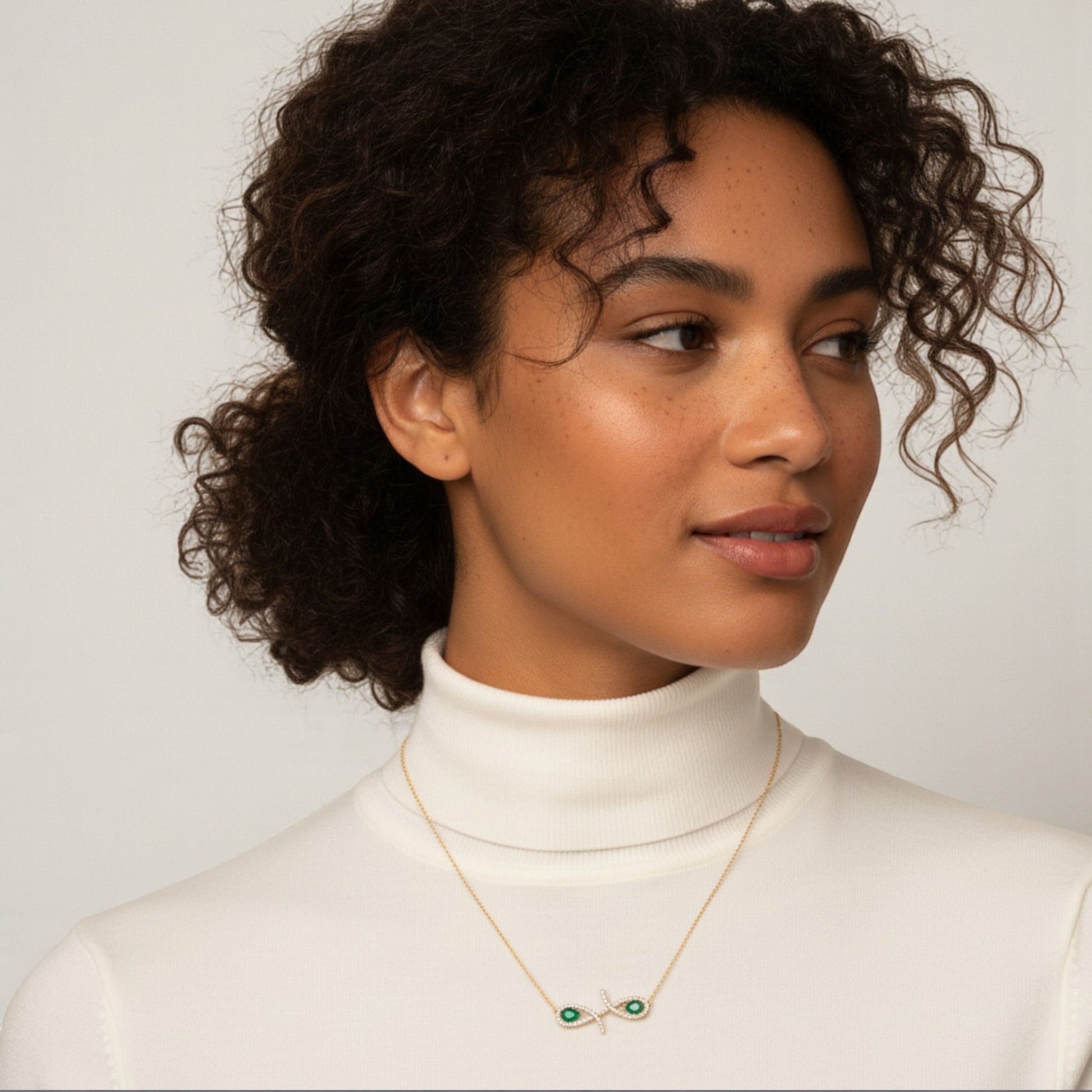 Woman wearing a white turtleneck and gold necklace with green gemstones against a plain background