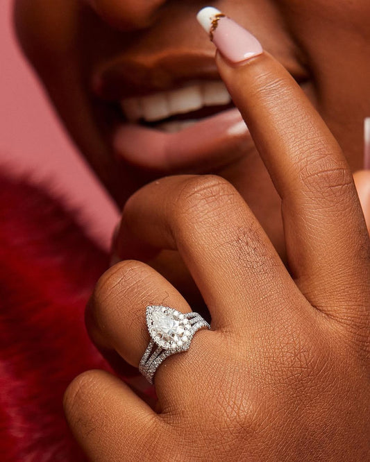 A person's hand displaying a pear-cut moissanite ring set in 18K gold, with a band adorned with natural diamonds.